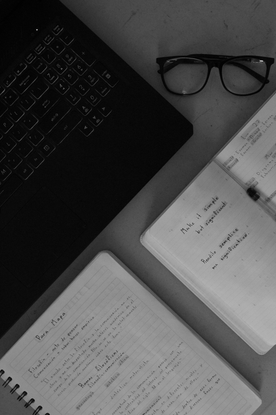Klawiatura laptopa, okulary i otwarte notatniki na biurku.