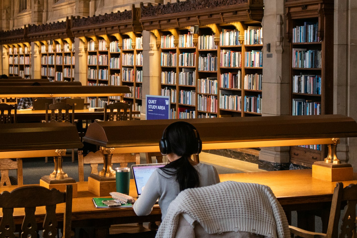 Osoba w słuchawkach pracuje na laptopie przy drewnianym biurku w bibliotece.